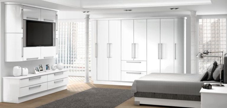 Bright bedroom with shiny white wardrobes & cupboards, a dark grey bed, mounted television & slim white pillars