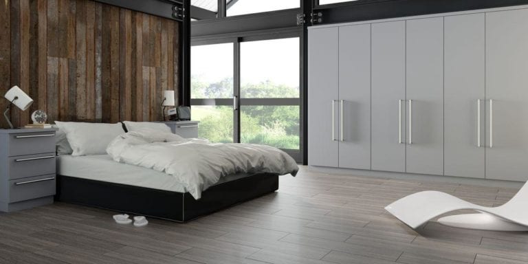 Bedroom with grey built in wardrobes, large window, a double bed that backs onto a dark wooden plank wall & flanked by grey bed side cabinets